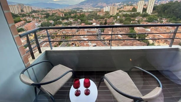 Varanda com duas cadeiras e uma mesa pequena, vista para um horizonte urbano com edifícios e montanhas.