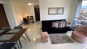 Uma sala de estar moderna com um sofá preto, uma mesa de jantar com cadeiras e uma pequena área de escritório com escrivaninha e cadeira.