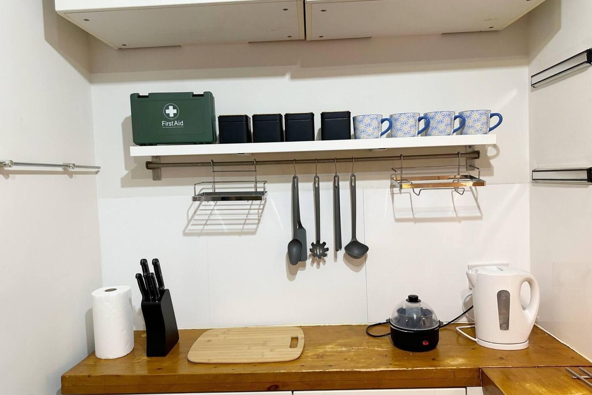 Une cuisine dotée d’un plan de travail en bois, d’une planche à découper, d’une bouilloire, d’une machine à café, d’un bloc à couteaux, d’un rouleau d’essuie-tout et d’ustensiles suspendus.