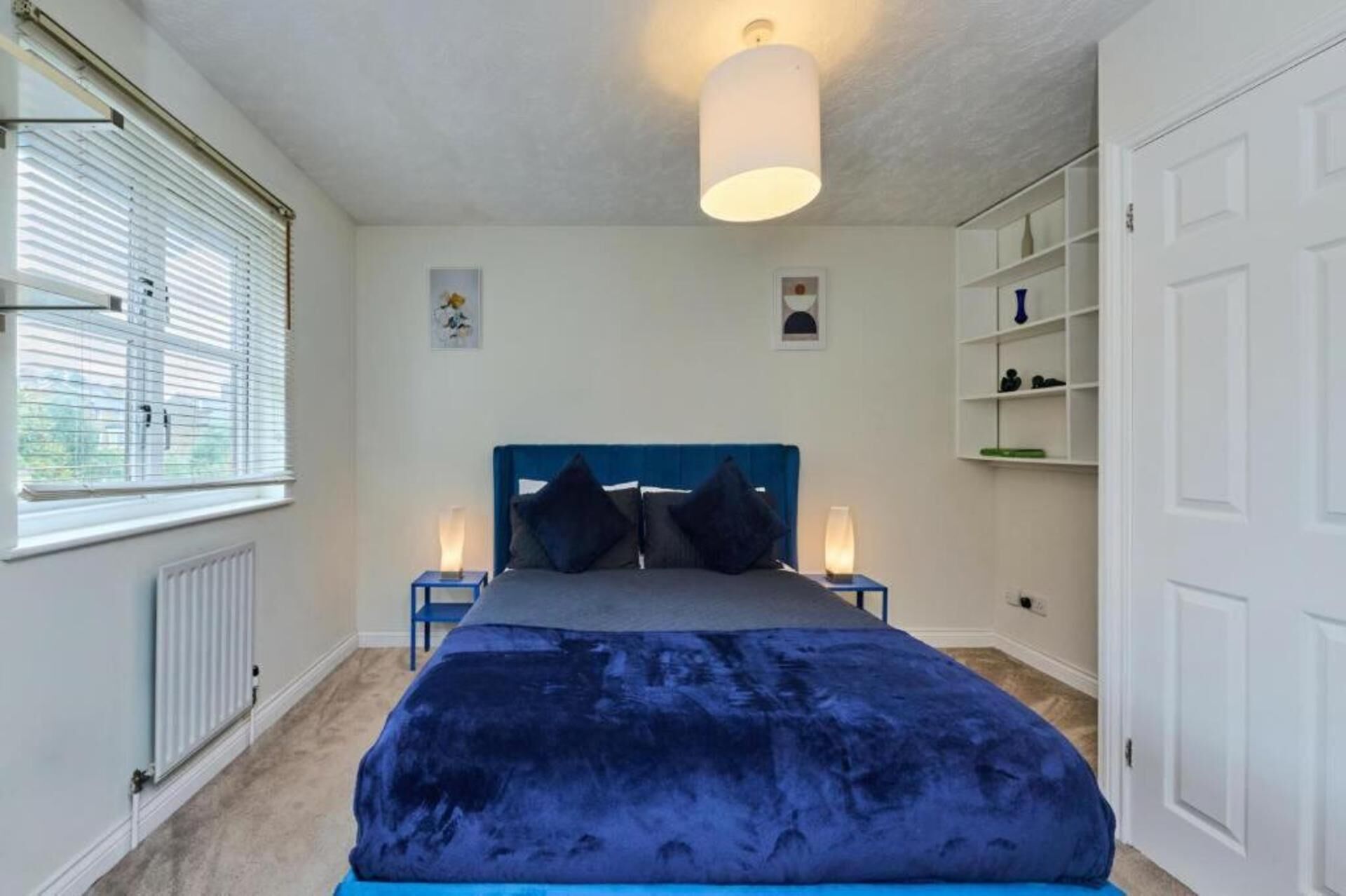Ein Schlafzimmer mit einem großen Bett, zwei Nachttischen mit Lampen, einem Heizkörper, einem Fenster mit Jalousien und einem weißen Kleiderschrank.