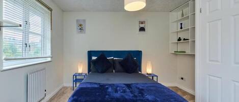 A bedroom with a large bed, two bedside tables with lamps, a radiator, a window with blinds, and a white wardrobe.