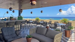 Zona de estar al aire libre con vista a la playa, con mesas redondas, sillas y luces esféricas colgantes.