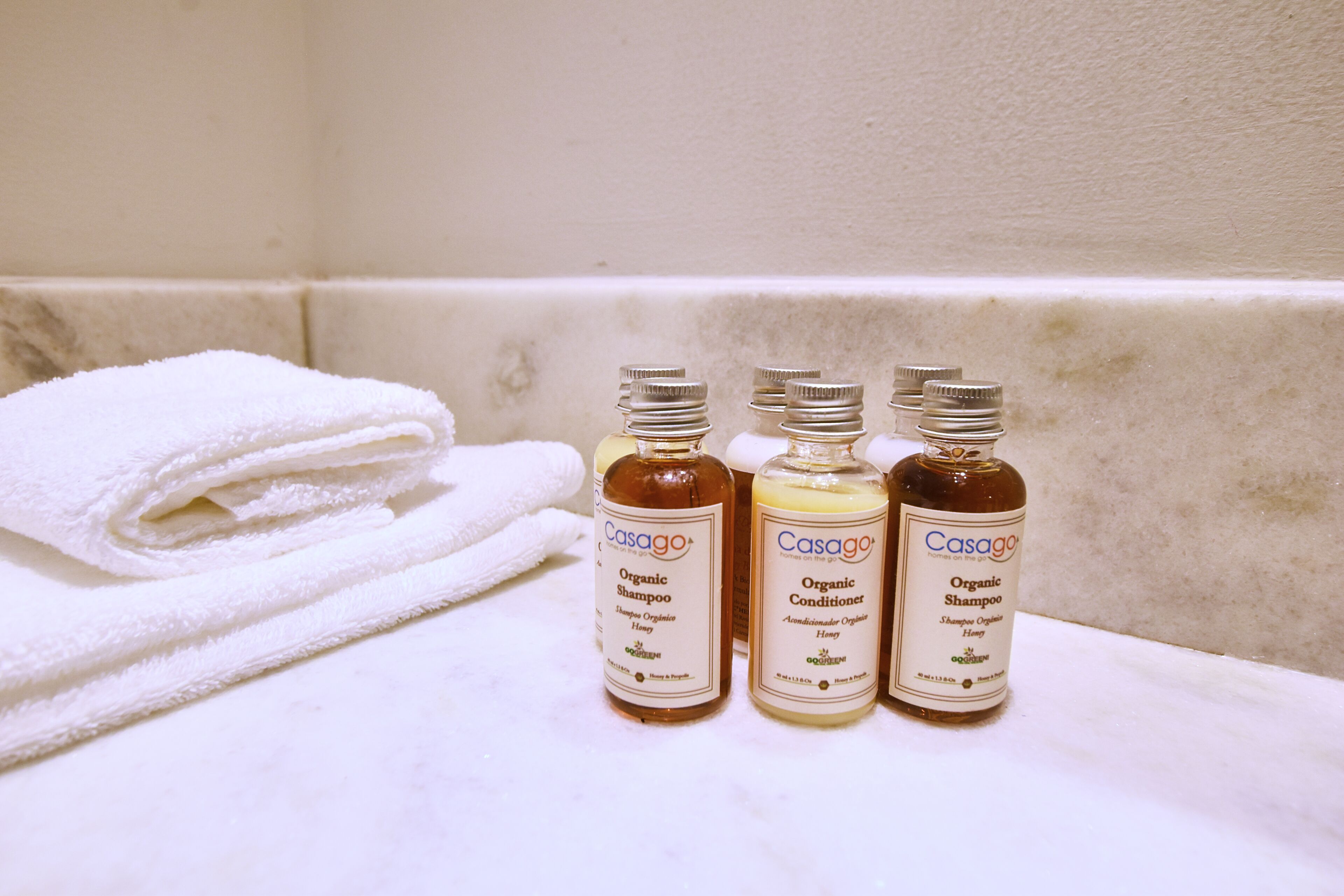 Trois bouteilles de shampoing et d’après-shampoing biologiques sont posées sur une surface carrelée blanche, avec une serviette blanche pliée en arrière-plan.