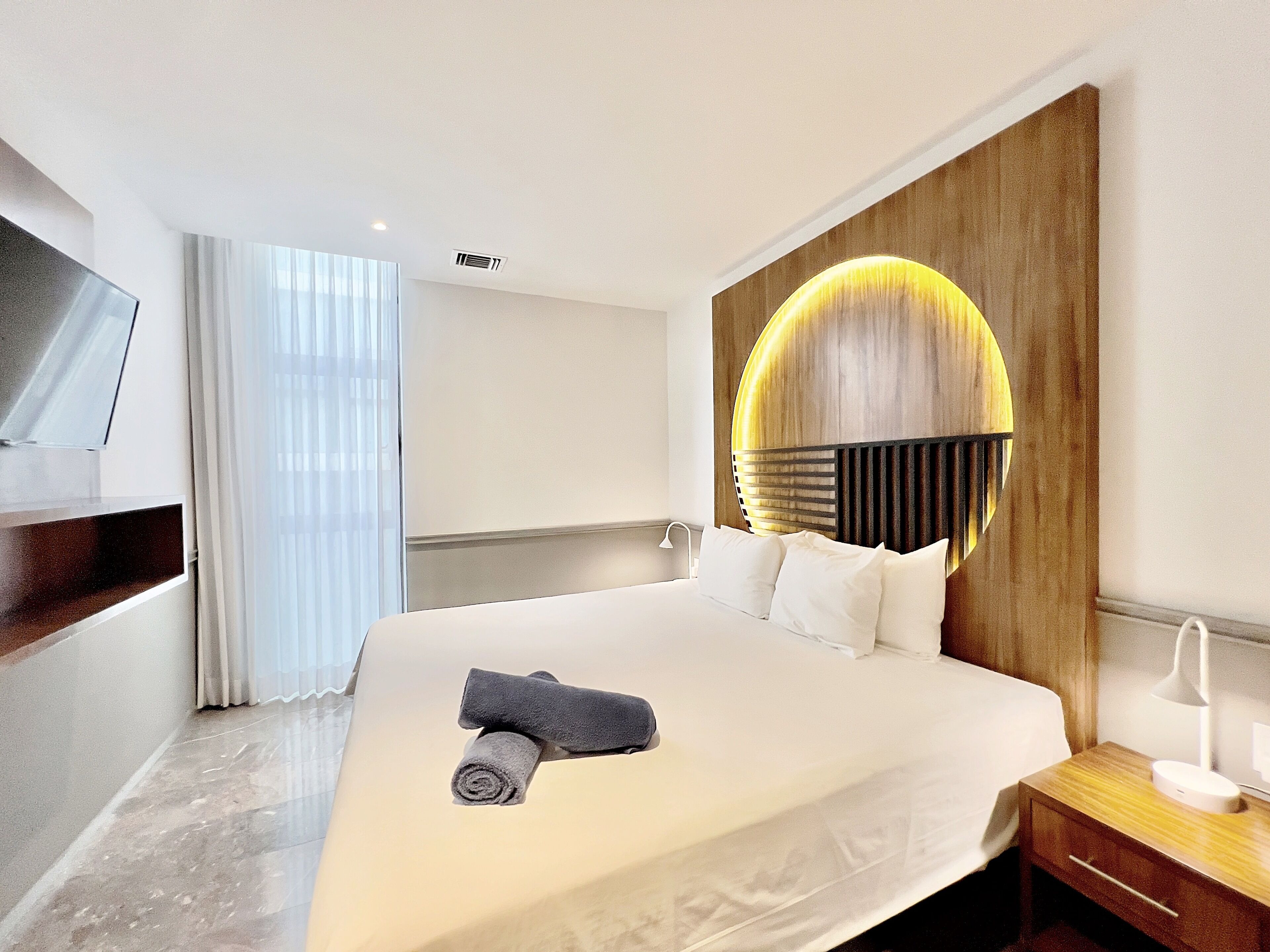 A modern hotel room with a large bed, a wooden headboard with a circular light, a bedside table with a lamp, and a TV mounted on the wall.