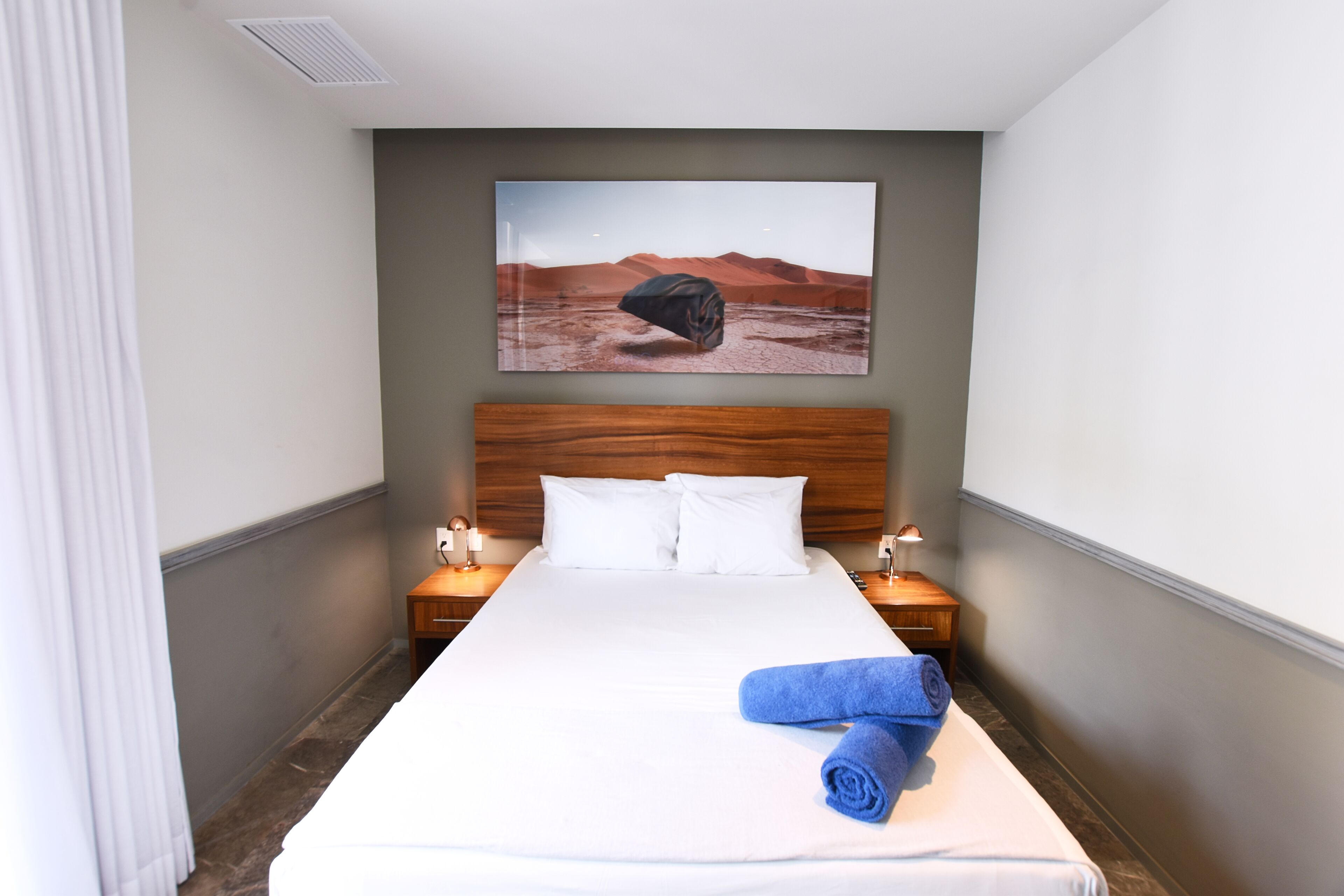 Una habitación de hotel con una cama grande, un cabecero de madera, dos mesitas de noche con lámparas y un cuadro de un paisaje desértico en la pared.
