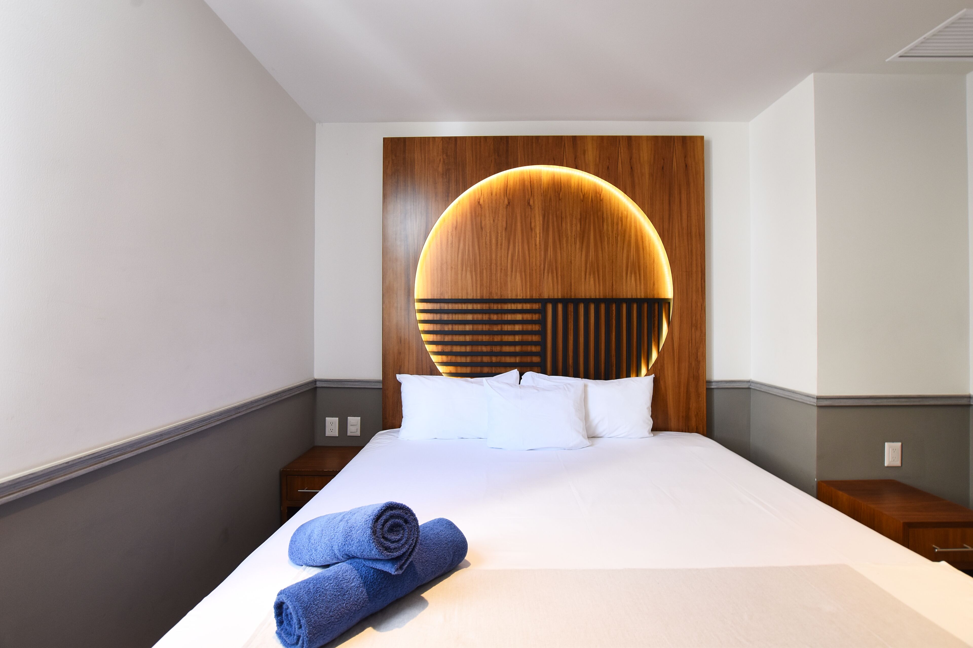 Une chambre d’hôtel avec un grand lit, une tête de lit en bois, du linge de lit blanc et une serviette bleue roulée posée sur le lit.