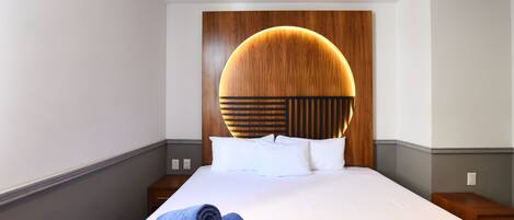 Une chambre d’hôtel avec un grand lit, une tête de lit en bois, du linge de lit blanc et une serviette bleue roulée posée sur le lit.