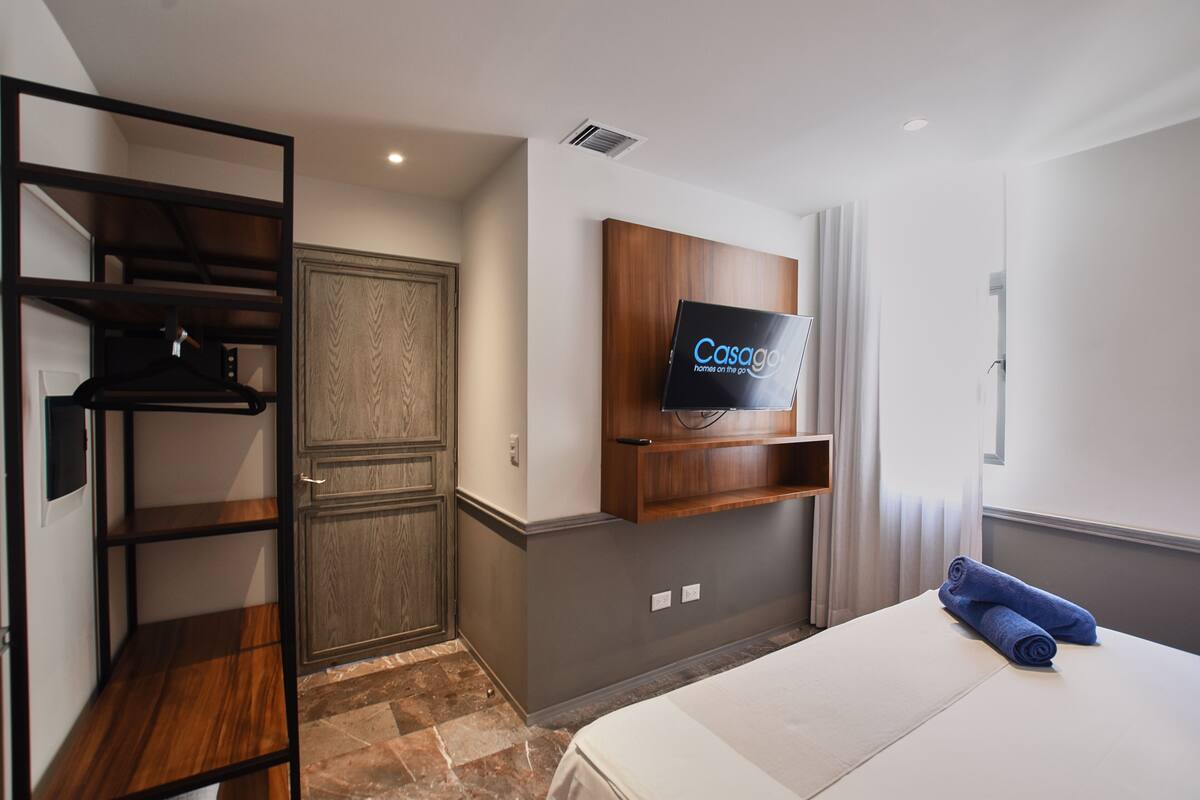 A hotel room with a bed, a TV mounted on the wall, a wooden wardrobe, and a built-in shelf.