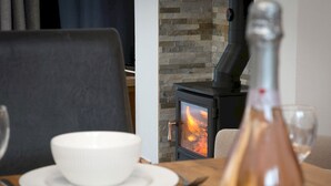 Una mesa de comedor con un tazón blanco, una copa de vino y una botella, una silla y una chimenea de piedra al fondo.