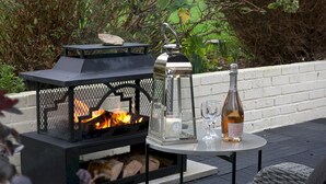Un área de estar al aire libre con una fogata integrada, un farol y una botella de vino sobre una mesa.