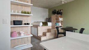 Una habitación moderna y luminosa con estanterías empotradas, un televisor de pantalla plana, una cama con manta verde y una zona de comedor con mesa y sillas.