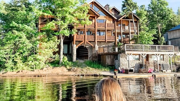 Een houten lodge met meerdere balkons met uitzicht op een kalm meer, omgeven door weelderige begroeiing.