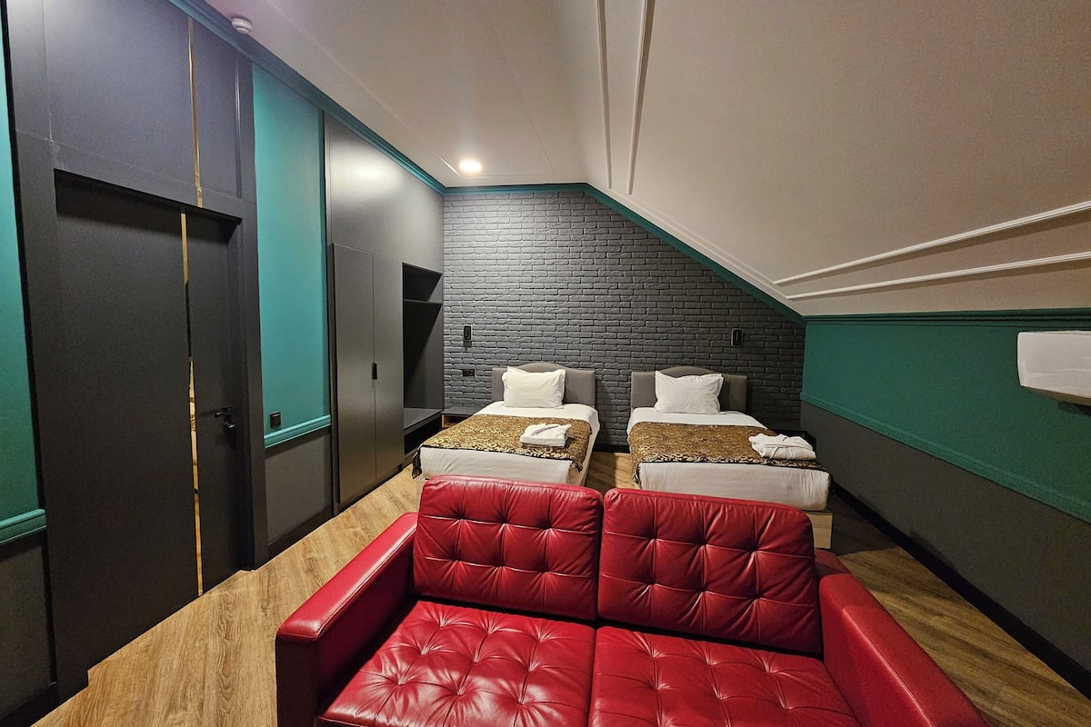 Ein Hotelzimmer mit zwei Betten, einem roten Ledersofa, einer Wand mit Backsteinoptik und einer dunklen Tür.