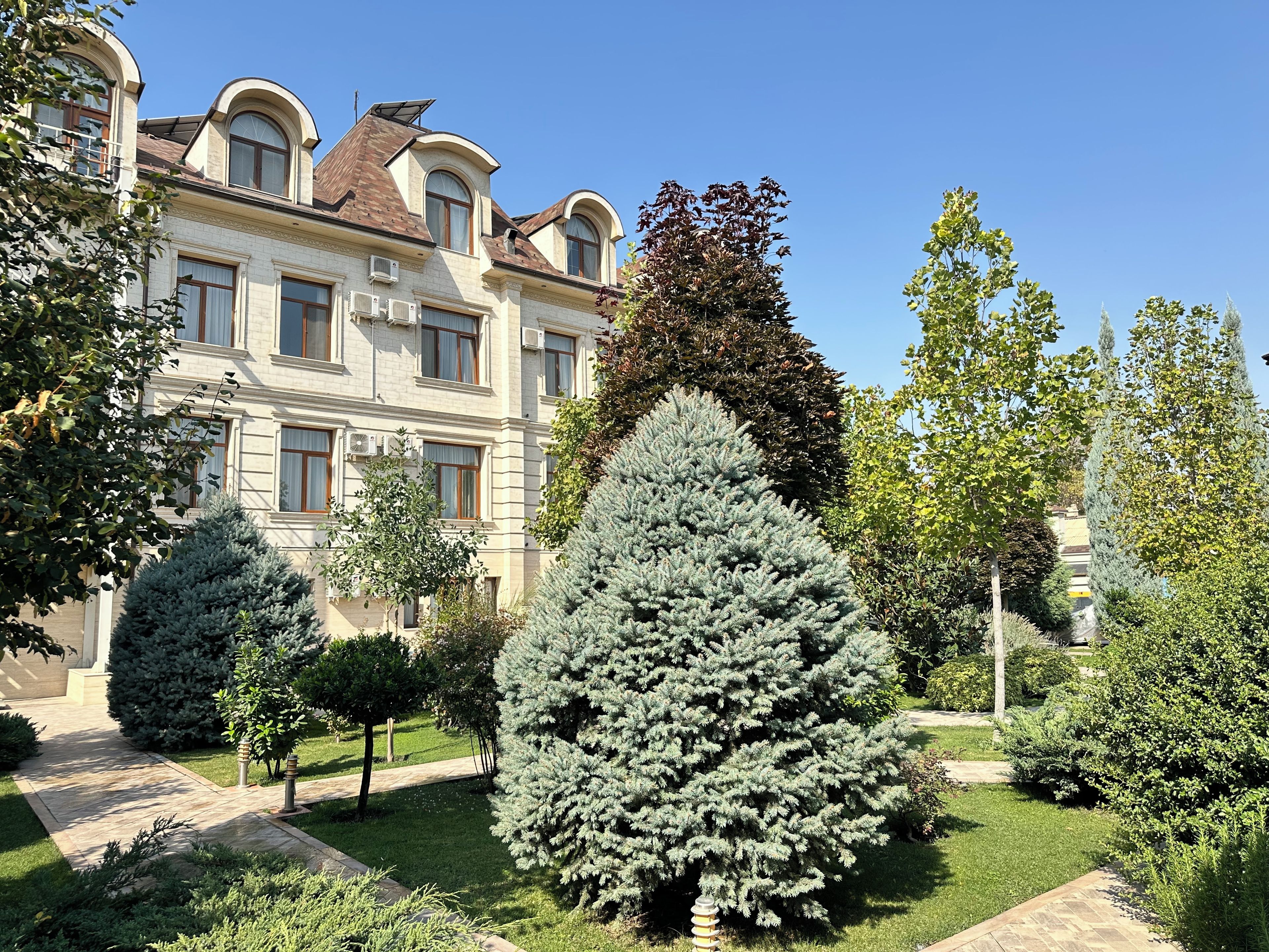 Un edificio grande de varios pisos con estilo arquitectónico clásico, rodeado de jardines cuidados y diferentes tipos de árboles.