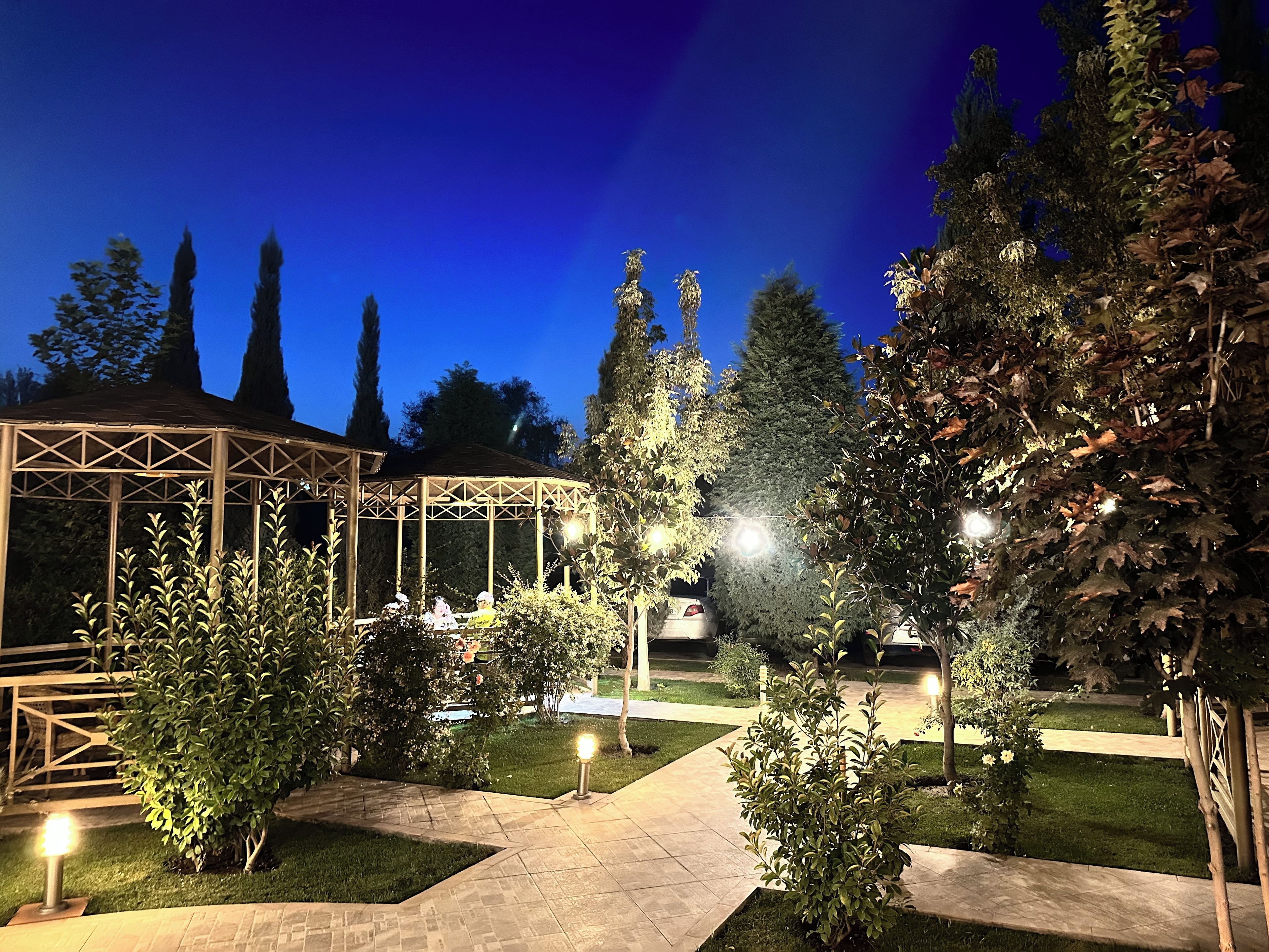 A well-lit garden area with a gazebo, pathways, and various trees.