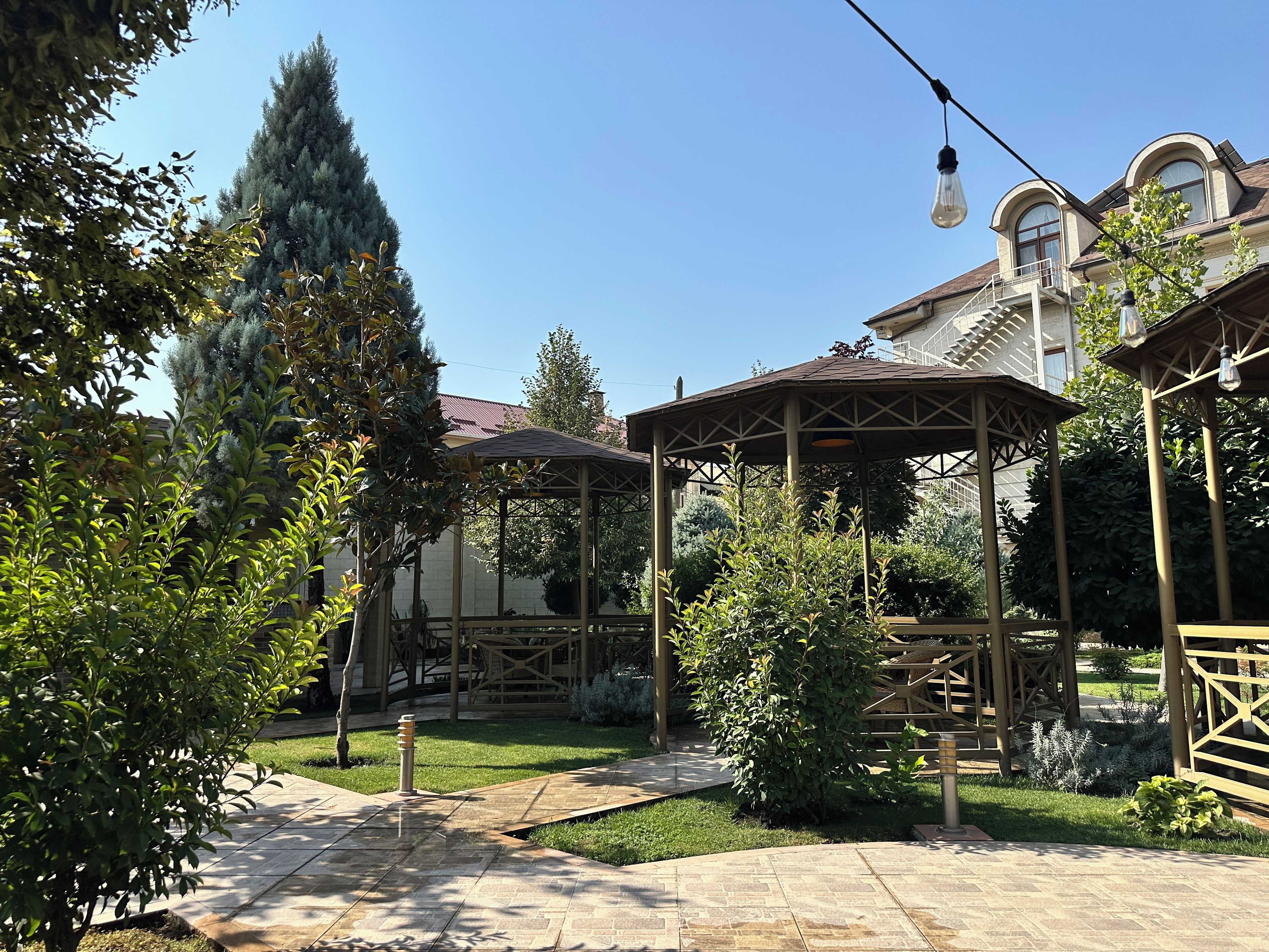 A gazebo with a tiled patio, surrounded by greenery and a building with a balcony in the background.