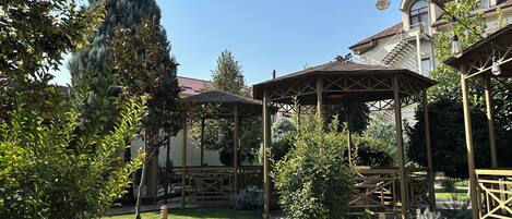 A gazebo with a tiled patio, surrounded by greenery and a building with a balcony in the background.