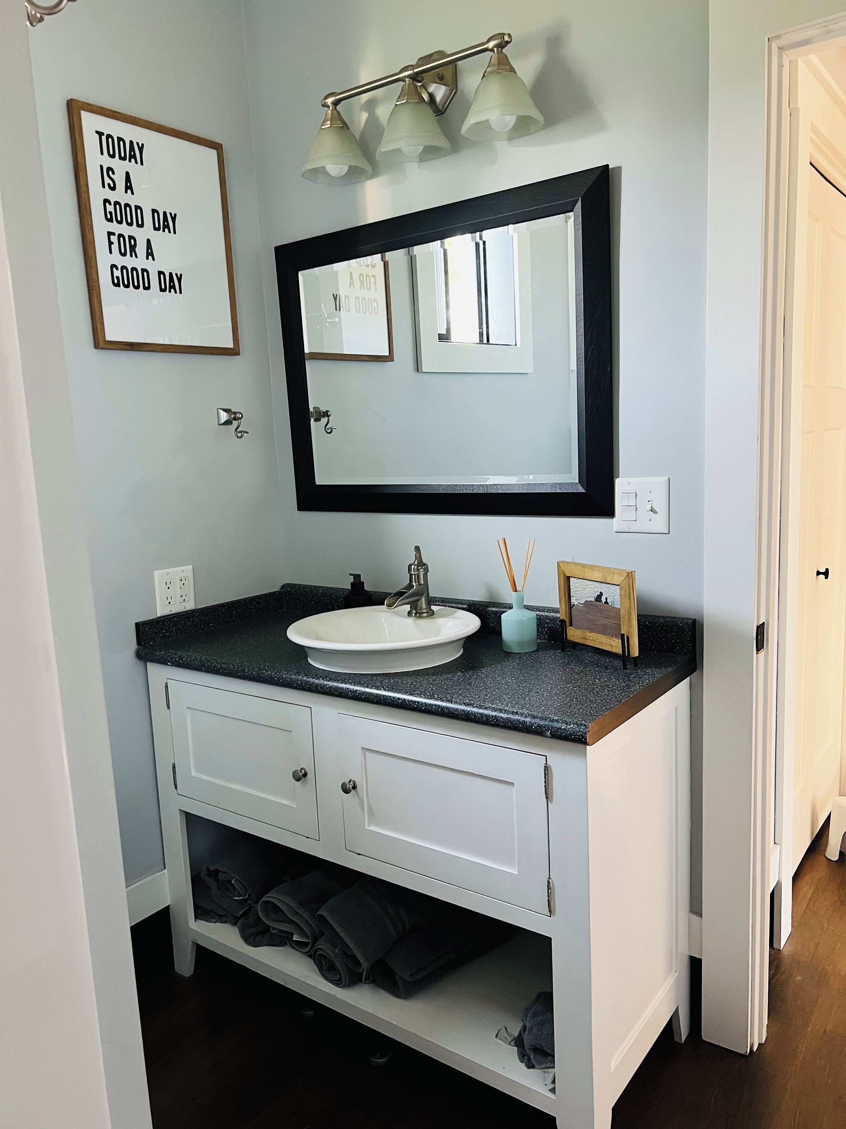 A bathroom with a white sink, black countertop, and a mirror with a black frame. There is a framed quote on the wall and a small picture frame on the countertop.