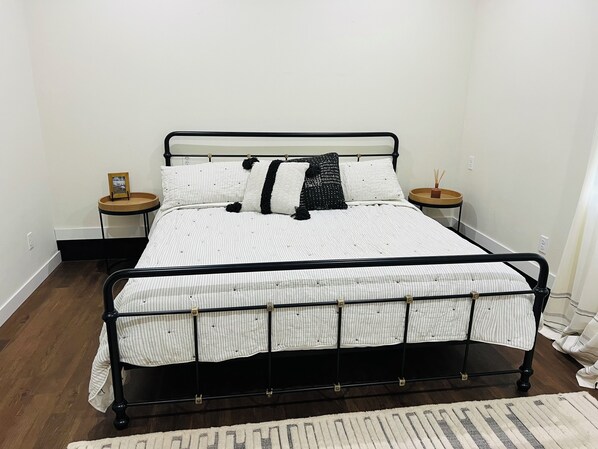 Una cama con estructura metálica, ropa de cama blanca a rayas y almohadas decorativas negras. Hay dos mesitas de noche, una con una lámpara pequeña y un marco de fotos. El cuarto tiene piso de madera y paredes blancas.