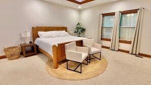 Quarto com cama de madeira, cadeiras brancas, mesa de cabeceira, abajur, planta e janela com cortinas.