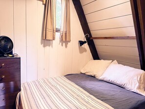 Cozy loft-style room with a bed, dresser, and fan, featuring a window with curtains.