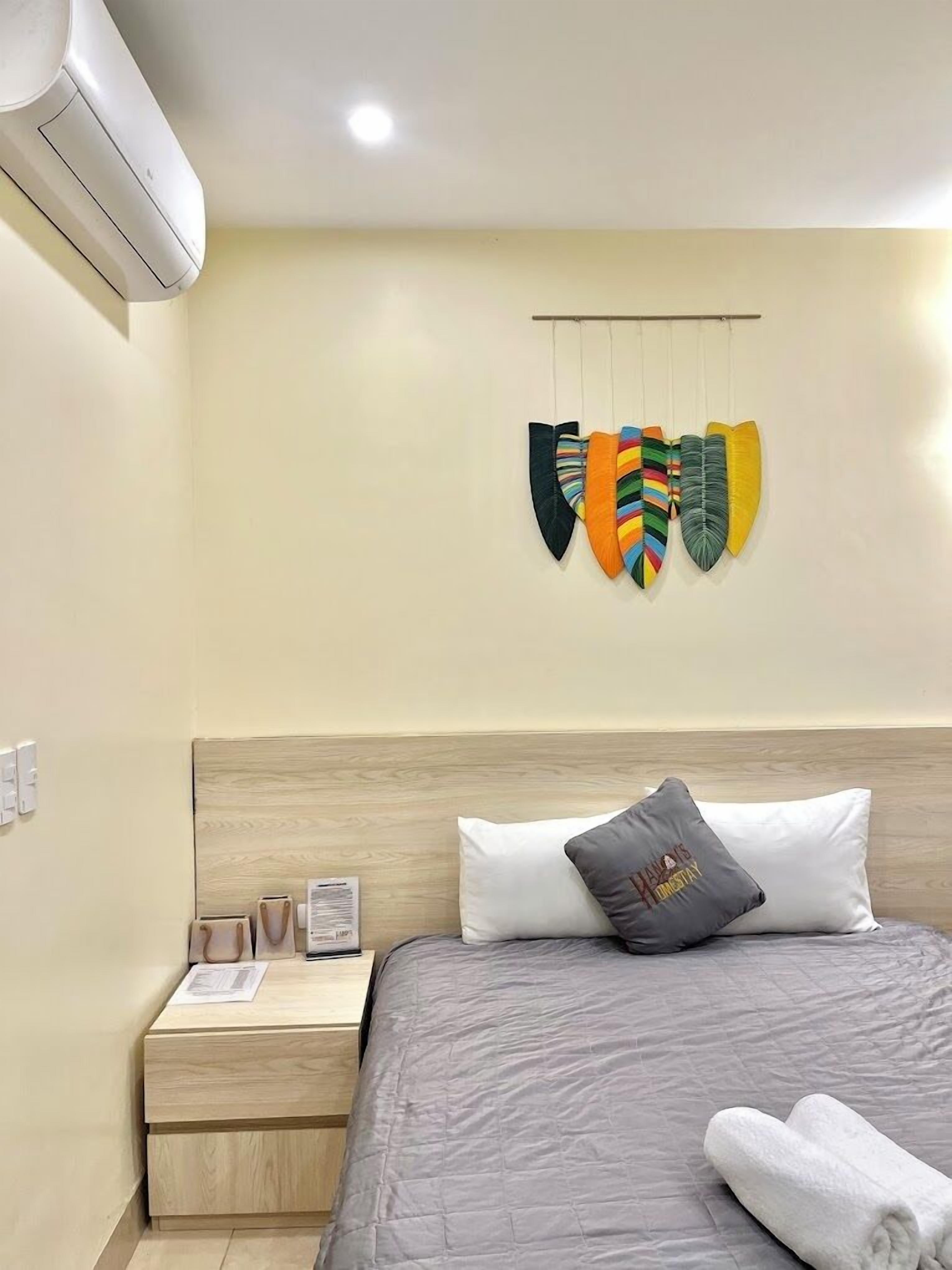 A hotel room with a bed, a nightstand, a wall-mounted air conditioner, and a wall decoration of colorful leaf-like objects.