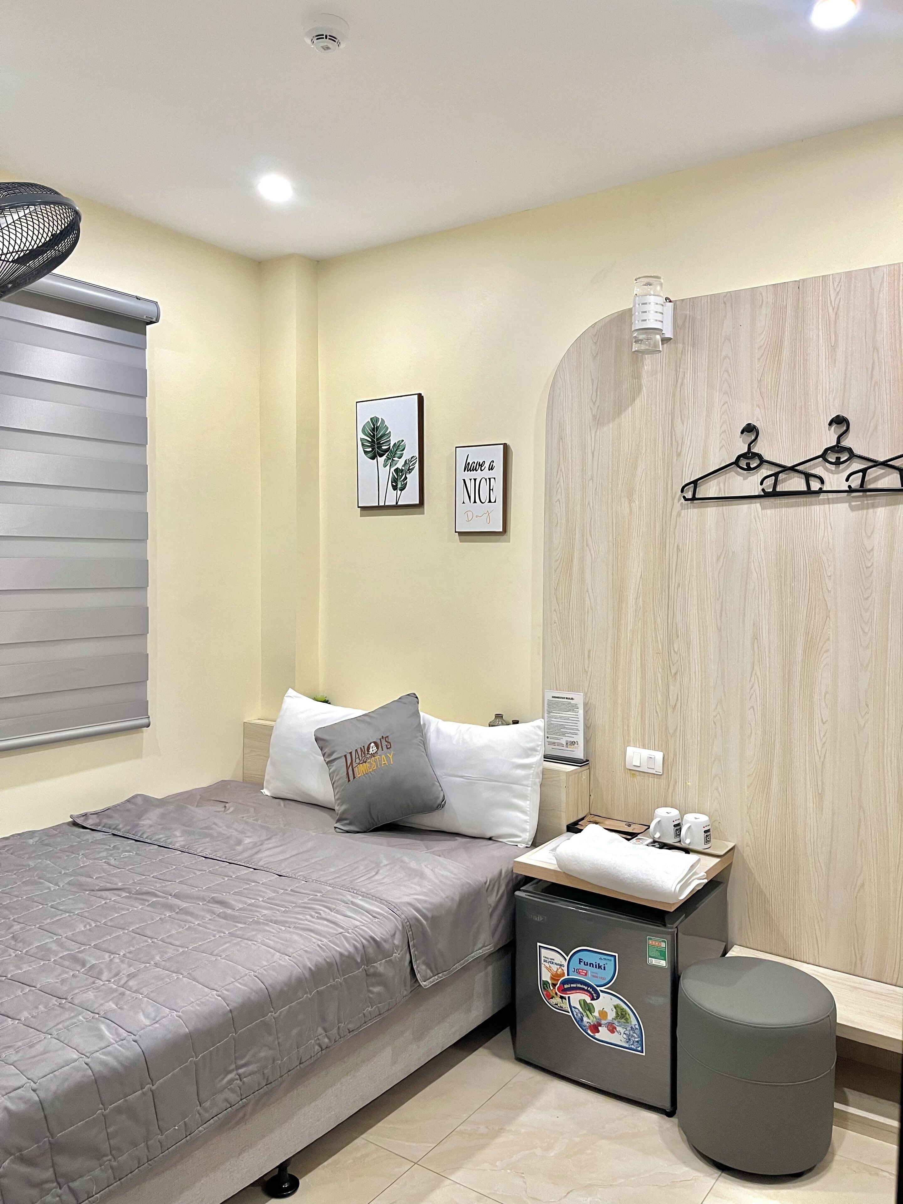 A neatly arranged hotel room with a bed, a bedside table with a lamp, a small stool, and a wall-mounted coat rack.
