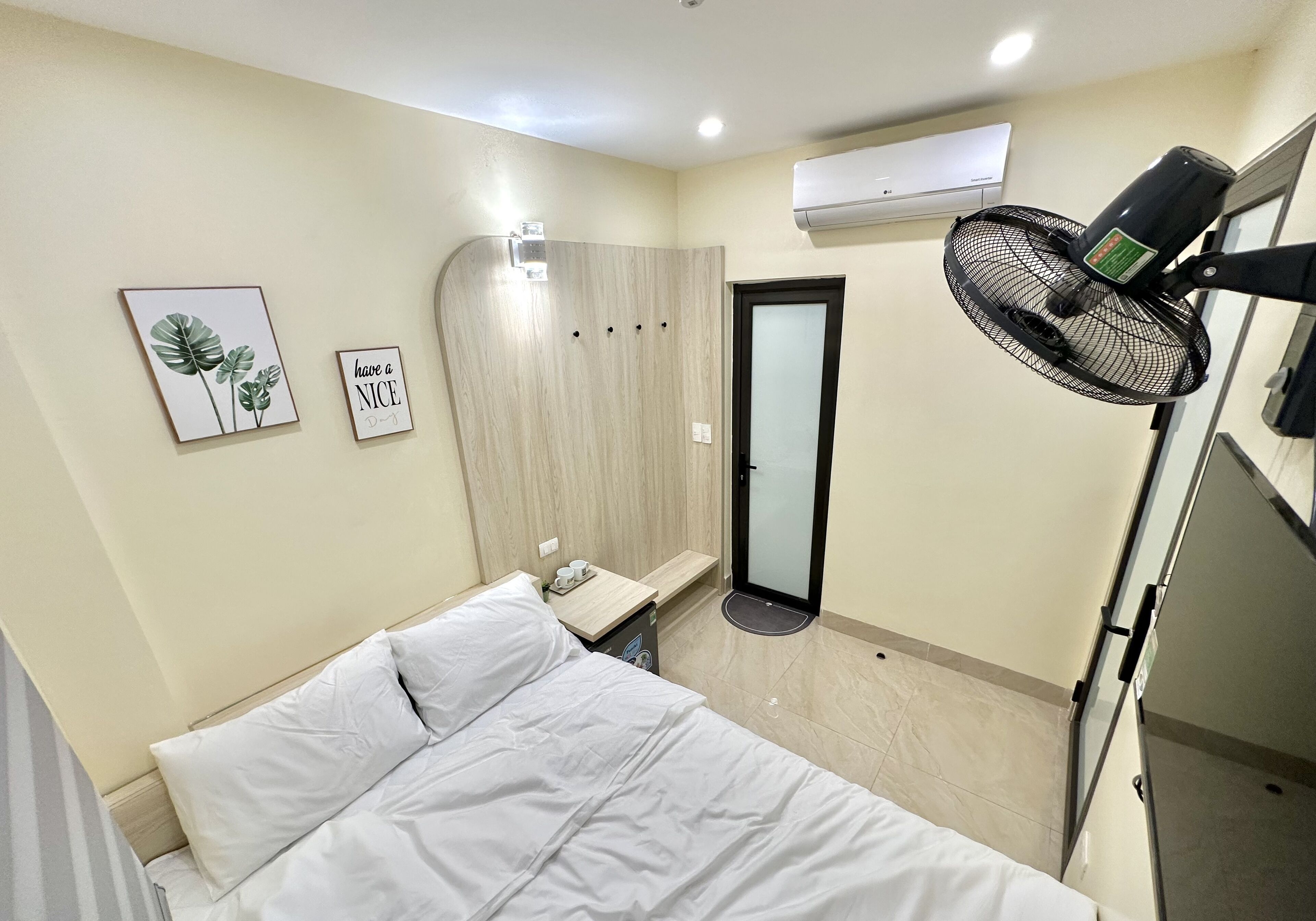 A hotel room with a bed, a bedside table, an air conditioning unit, a wall-mounted fan, and a door with a glass panel.