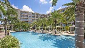 Une grande piscine entourée de palmiers et de chaises longues, avec un immeuble d’hôtel à plusieurs étages en arrière-plan.