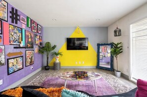 Experience the vibrant atmosphere of this eclectic living room, featuring a colorful wall of artwork, a bold accent wall, and a cozy seating area with a flat-screen TV.