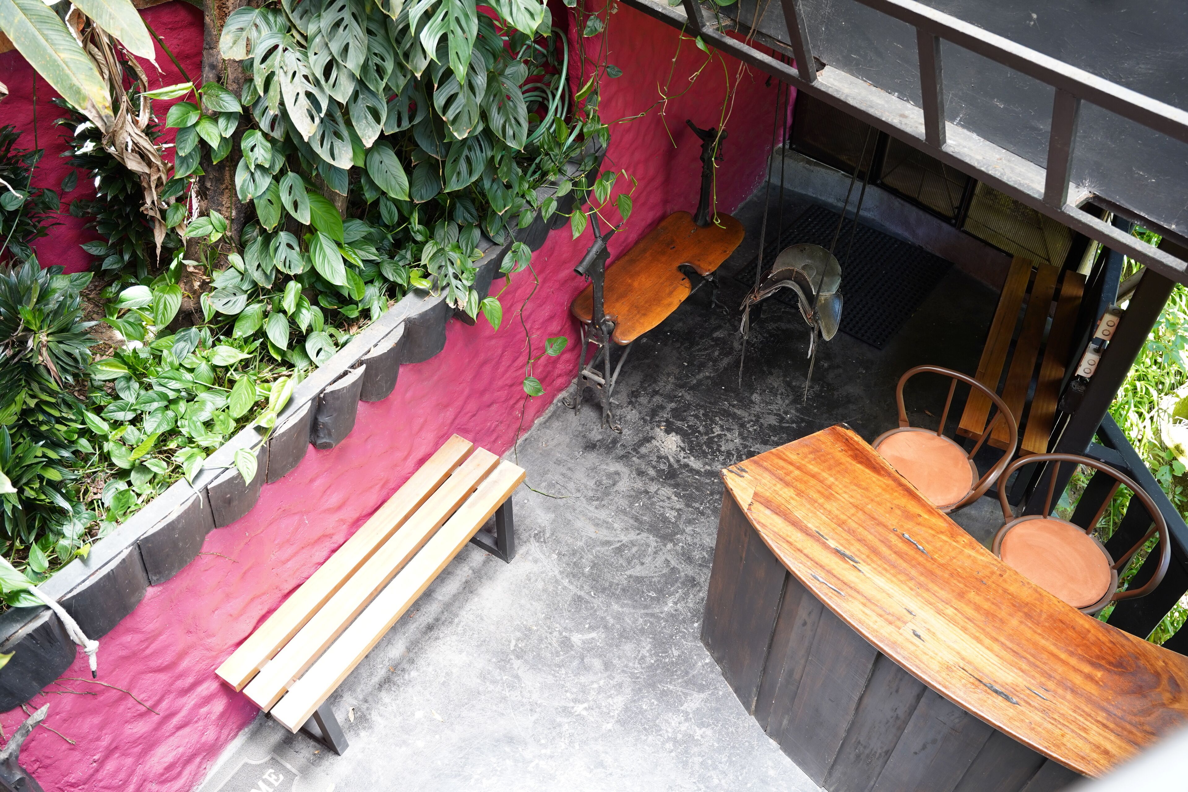 A wooden bench and a wooden table with chairs in an outdoor area with a red wall and green plants.