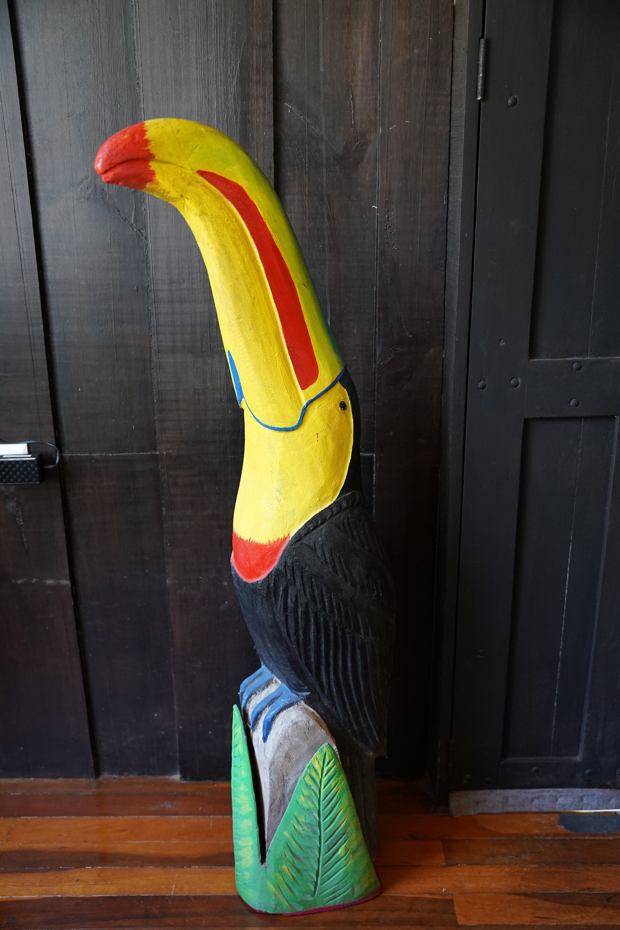 A wooden sculpture of a toucan with a large beak, standing on a base with a leaf design.