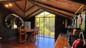 A rustic wooden interior with a dining table, chairs, a refrigerator, and a large window offering a view of greenery. - TreeTop House Getaway, Fairy Magic Rustic Elegance in the Cloud Forest, 3 Levels (Monteverde)