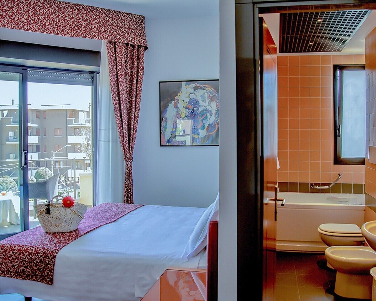 A hotel room with a bed, a window with curtains, a painting on the wall, and a bathroom with a bathtub and toilet.