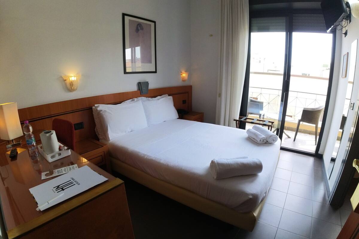 Une chambre d’hôtel avec un lit bien fait, une table de chevet avec une lampe, un tableau encadré au mur et une vue sur le balcon extérieur.