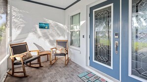 Un porche avec des fauteuils à bascule et une petite table, une porte bleue avec un panneau en verre et un paillasson.