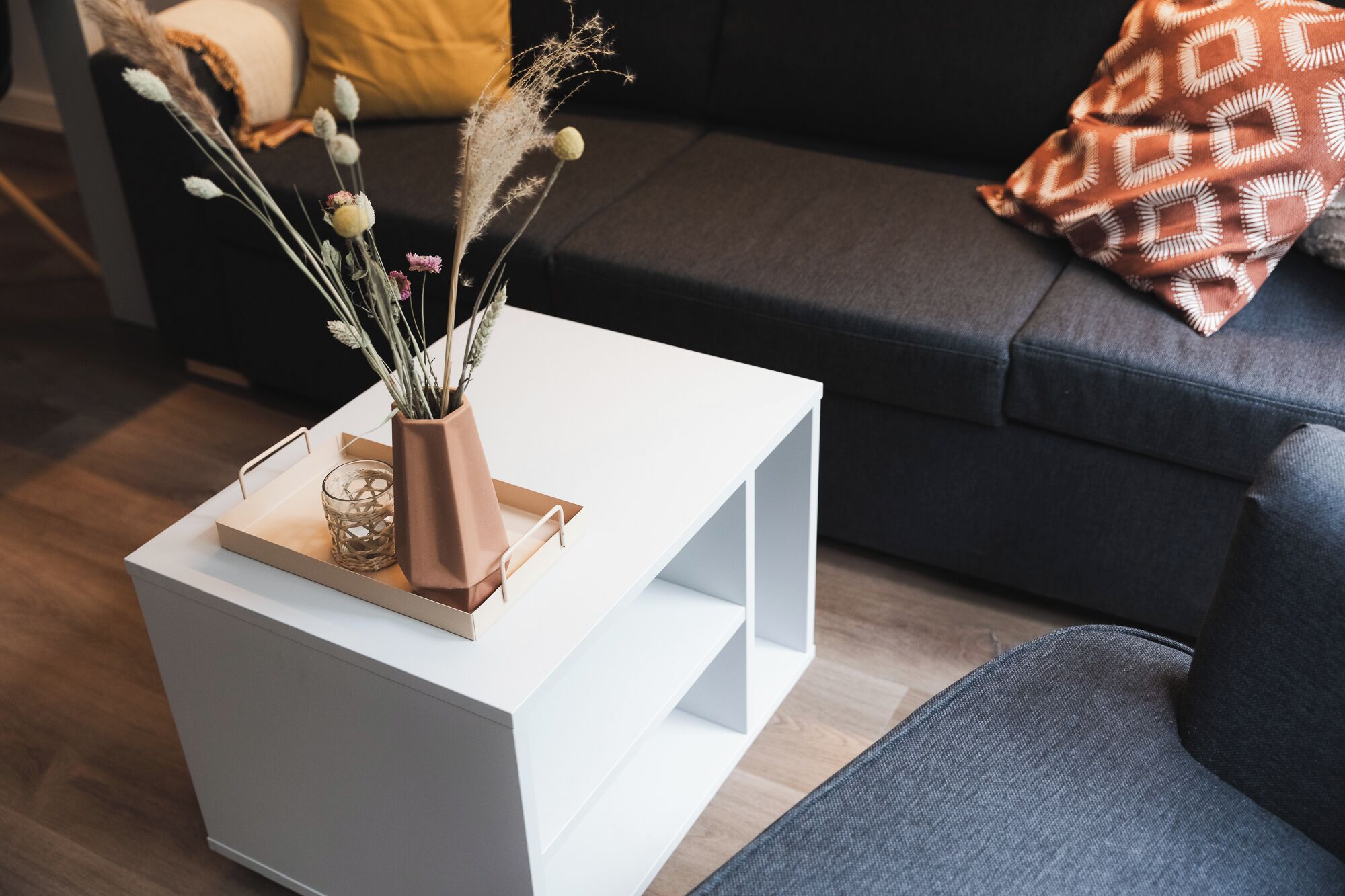 Een witte bijzettafel met een vaas gedroogde bloemen en een glas erop, geplaatst voor een donkergrijze bank met een kussen met dessin.