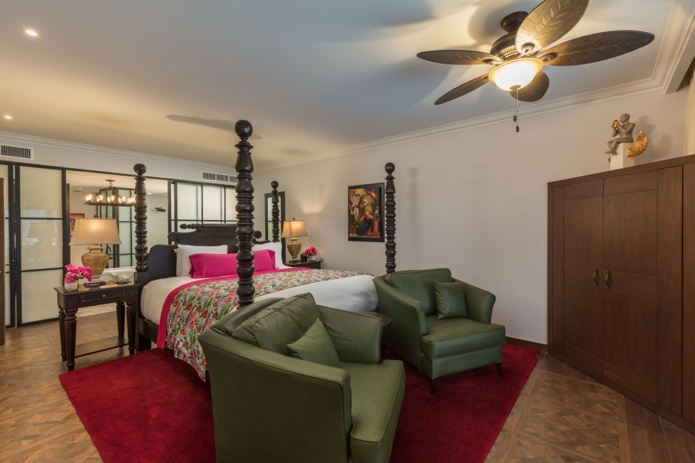 A four-poster bed with a floral bedspread, two green armchairs, a wooden wardrobe, and a ceiling fan.