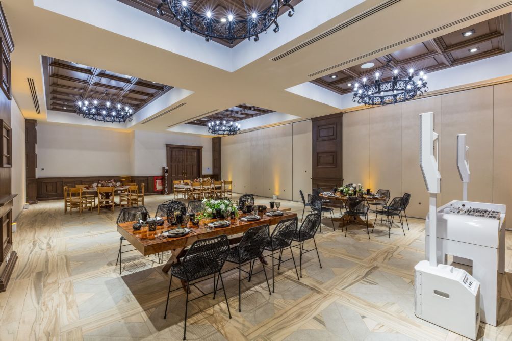 A spacious dining area with multiple tables and chairs, a chandelier, and a modern kitchen area with a white appliance.