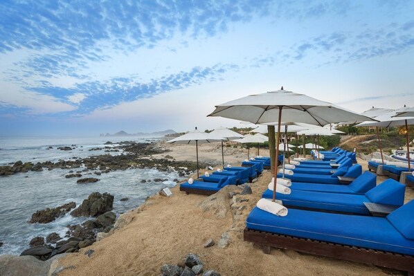Un espace de détente aménagé sur la plage, avec des fauteuils bleus dotés de coussins et des parasols blancs, offrant une vue sur une côte rocheuse et l’océan.