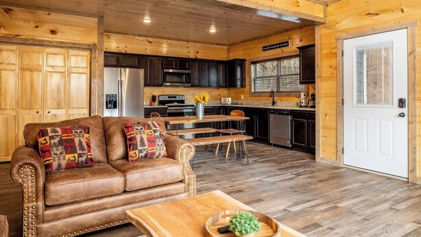 A cozy cabin interior with a leather sofa, wooden dining table, and a kitchen area with dark cabinets and a white door. - Smokies Cabin: Dog-Friendly, Hot Tub & Arcade Game (Sevierville)