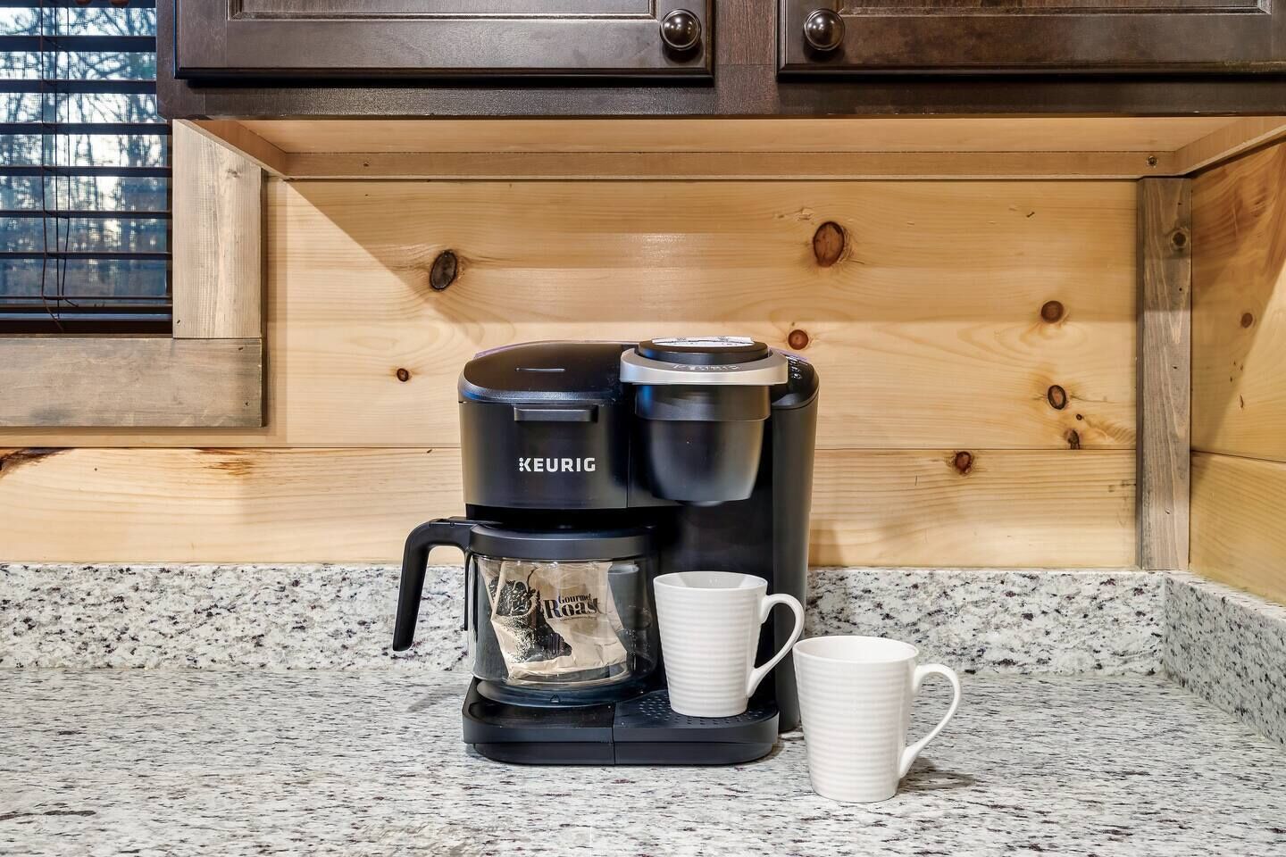 Une machine à café Keurig posée sur un comptoir en granit, avec deux tasses blanches à côté.