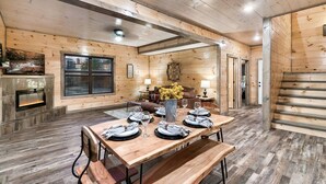 A cozy log cabin interior with a dining area, fireplace, and a staircase. - Smokies Cabin: Dog-Friendly, Hot Tub & Arcade Game (Sevierville)