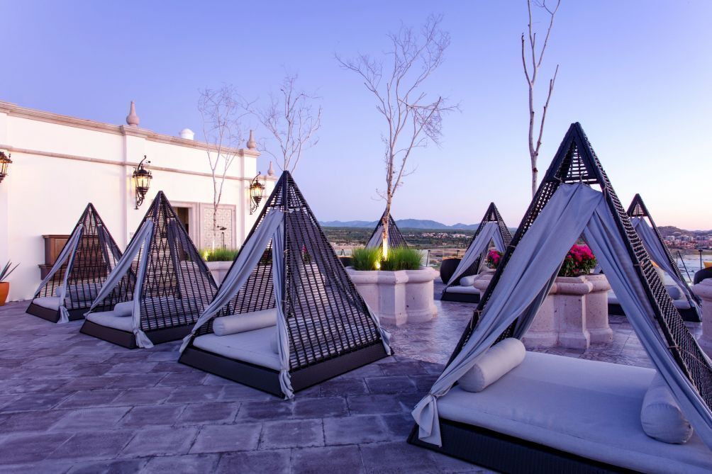 Outdoor seating areas with cushioned chairs and tables, arranged on a stone patio with a view of a building and landscape in the background.