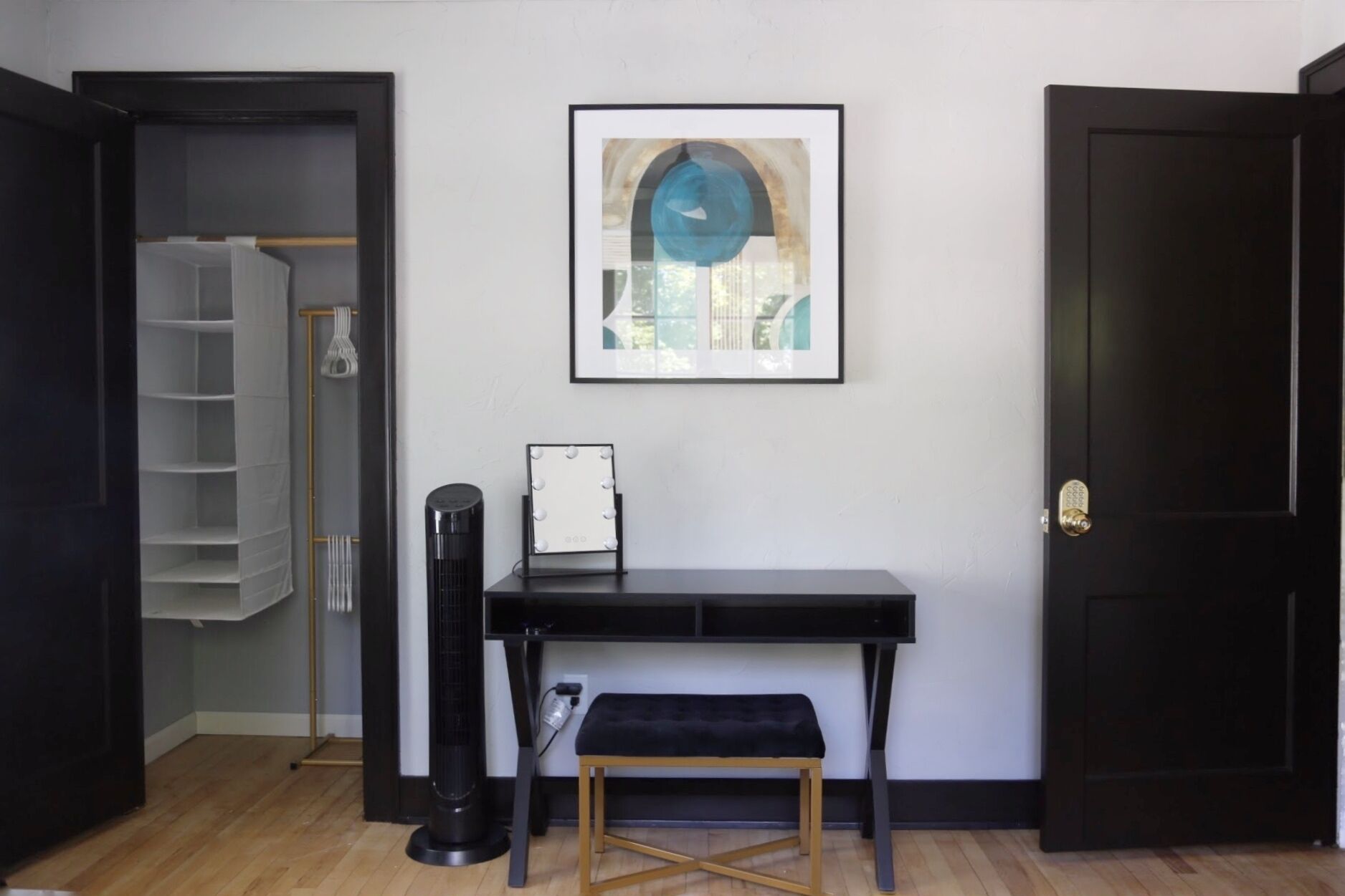 A room with a black desk, a bench, a standing fan, and a framed picture on the wall.