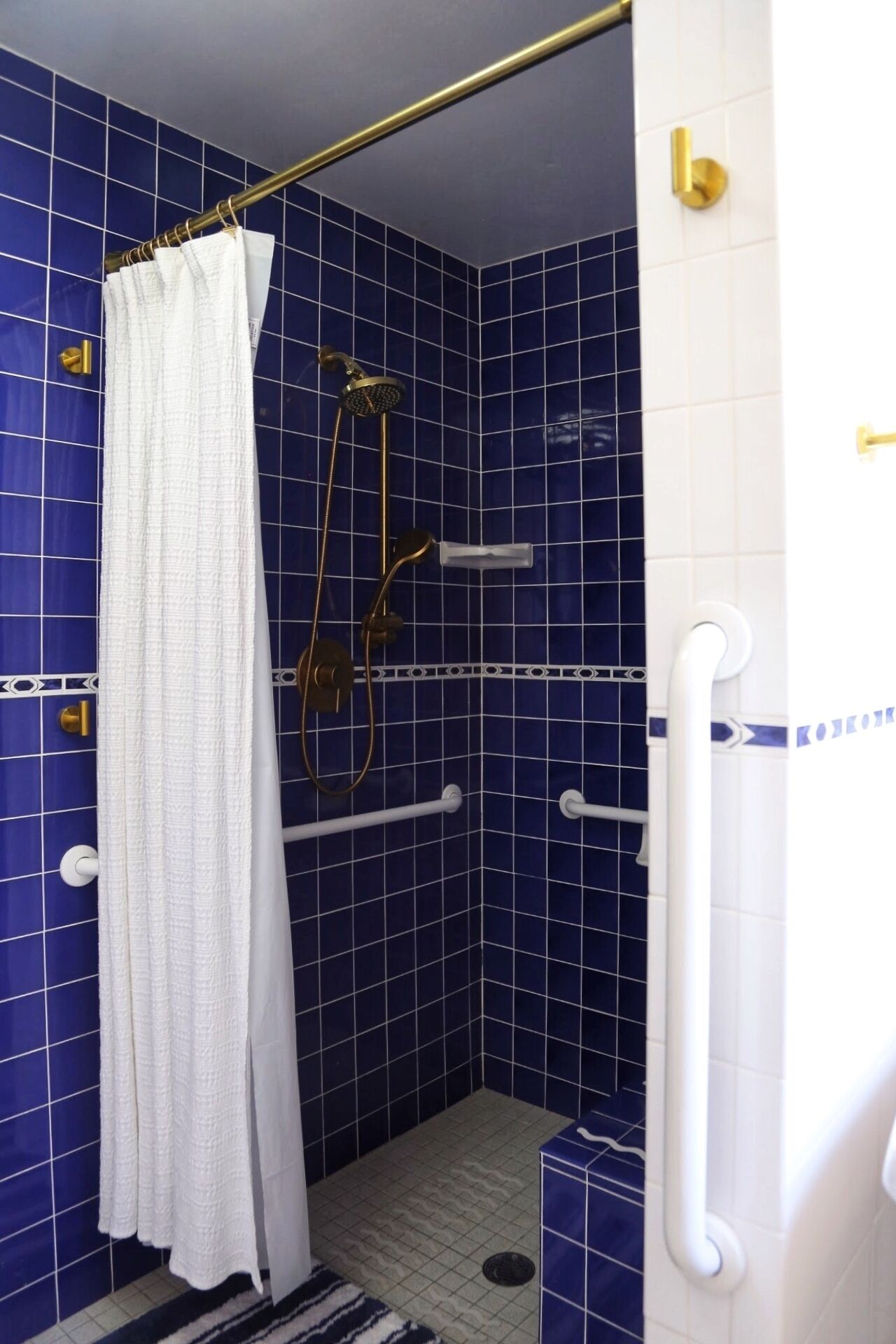 A small, blue-tiled shower stall with a white shower curtain, a handheld showerhead, and a handheld massage wand.
