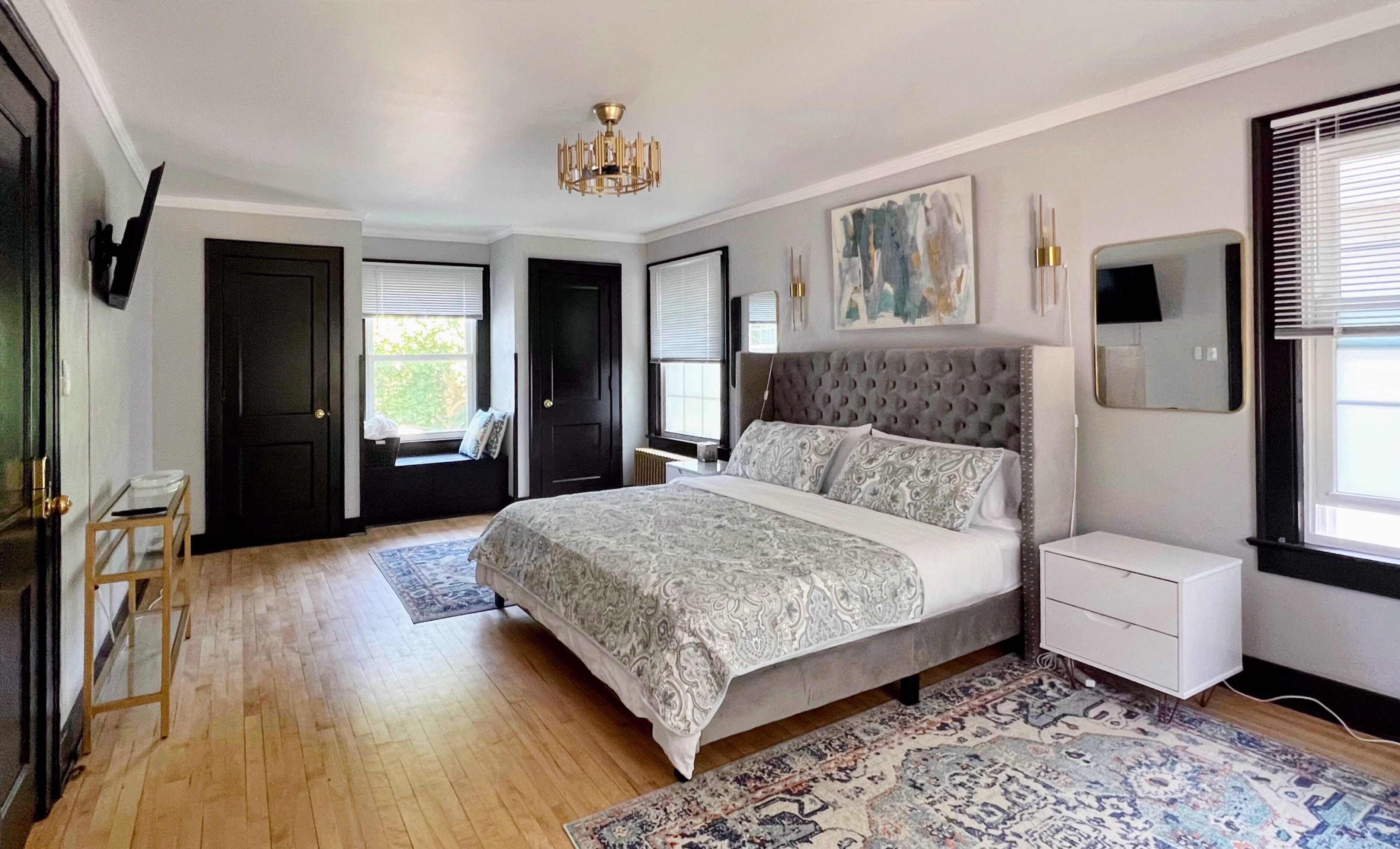 Une chambre à coucher avec un grand lit, une table de chevet, une commode, un lustre, une fenêtre avec des stores et un tableau au mur.