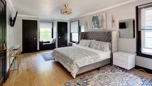 A bedroom with a large bed, a bedside table, a nightstand, a chandelier, a window with blinds, and a painting on the wall. - Riverfront Getaway (Two Rivers)