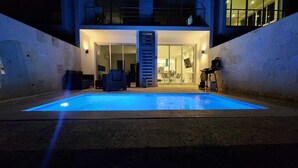 Una piscina con cerramiento de vidrio, un área de estar techada al aire libre y un edificio con balcón al fondo.