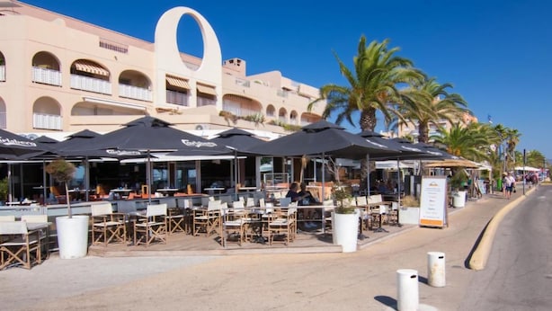 An outdoor café with tables and chairs, umbrellas, and a building with balconies in the background. - Nice studio with terrace port de hyeres (Hyères)